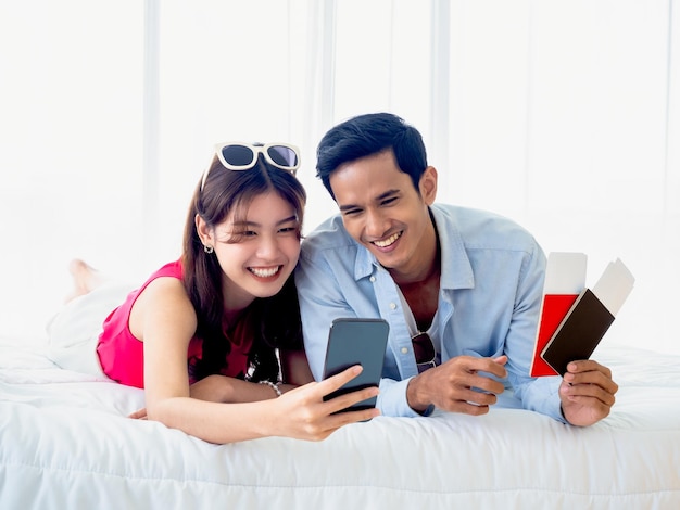 Heureux couple asiatique utilisant un téléphone portable Jeune homme et femme décontractés se sentent heureux de sourire et de regarder l'écran du smartphone tout en détenant un passeport pour la réservation de vol sur un lit blanc dans une chambre lumineuse