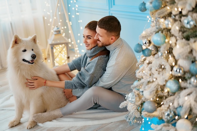 Heureux couple assis et étreignant près de l'arbre de Noël à la maison foyer vêtements et chandails