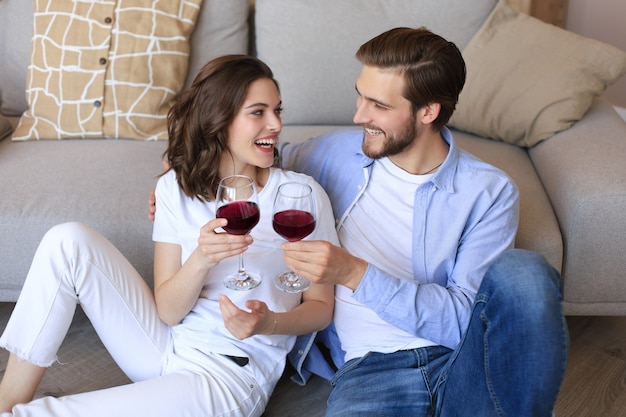 Heureux couple assis, relaxant sur le sol dans le salon, buvant du vin rouge. Un jeune mari et une femme souriants se reposent à la maison et profitent d'un rendez-vous romantique le week-end en famille ensemble.