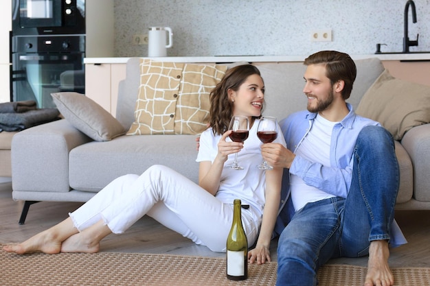 Heureux couple assis se détendre sur le sol dans le salon en buvant du vin rouge Souriant jeune mari et femme se reposent à la maison profiter d'un rendez-vous romantique le week-end en famille ensemble