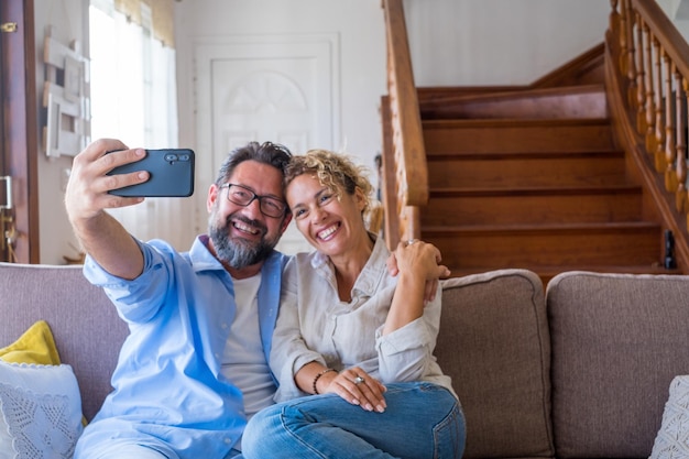 Heureux couple caucasien prenant selfie assis sur un canapé à l'aide d'un appareil photo de téléphone portable à la maison Joyeuse femme et mari prenant un autoportrait sur smartphone