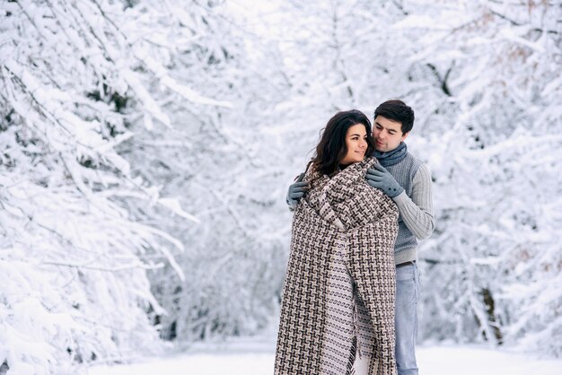 Heureux couple charmant recouvert d'une couverture chaude et marchant dans un parc enneigé