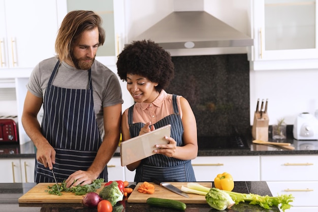 Heureux couple diversifié dans la cuisine utilisant une tablette et préparant la nourriture