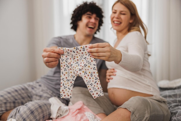 Heureux couple enceinte montrant des vêtements de bébé tout en se relaxant sur un lit dans la chambre à la maison.