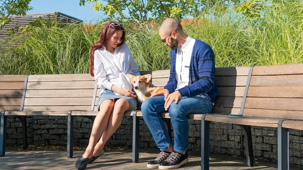 L'heureux couple est assis sur le banc dans le parc avec un petit chien mignon chiot corgi Pembroke