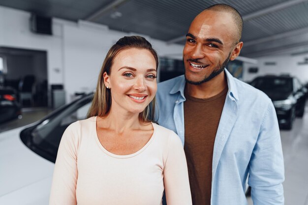 Heureux couple de femme caucasienne et homme afro-américain debout près de leur nouvelle voiture à l'intérieur du salon de l'automobile