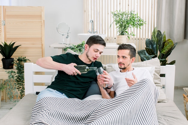 Heureux couple gay allongé sur le lit à la maison, un homme verse du vin dans des verres. Concept de couple gay