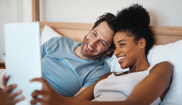 Photo heureux couple interracial relaxant et insouciant sur tablette numérique souriant et prenant un selfie au lit mari et femme se réveillant et lisant les nouvelles des médias sociaux sur écran tactile tout en se relaxant à la maison