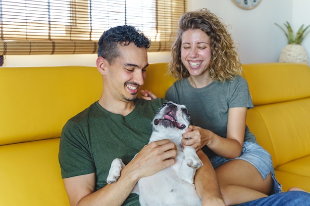 Heureux couple jouant avec un chien à la maison. Vue horizontale du couple caressant l'animal de compagnie de bouledogue sur le canapé.