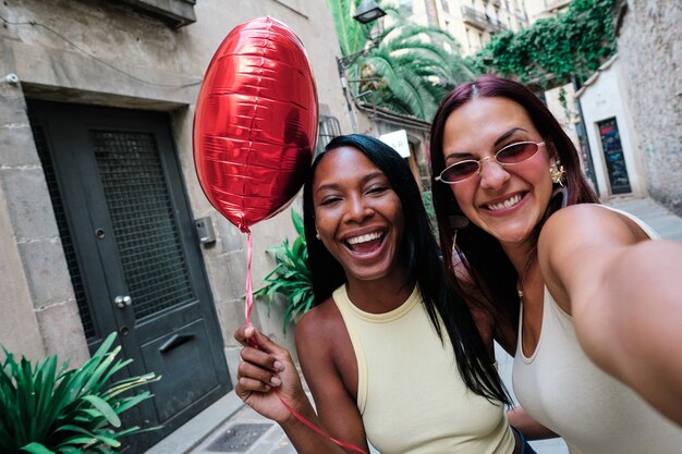 Photo heureux couple lesbien avec un ballon rouge en forme de coeur tout en prenant un selfie ensemble à l'extérieur dans la rue. notion lgbt.