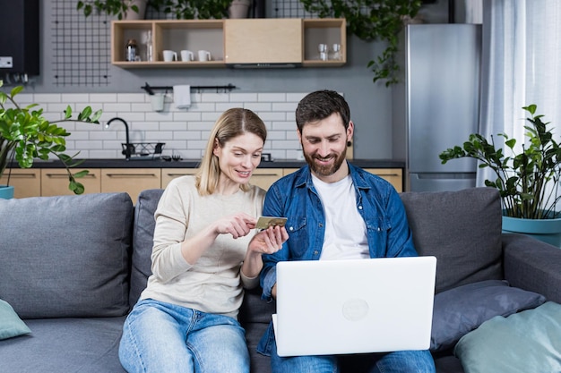 Heureux couple marié homme et femme assis à la maison sur le canapé à l'aide d'un ordinateur portable pour faire des achats en ligne détenant une carte bancaire de crédit