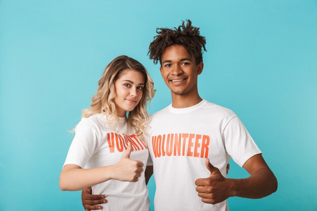 Heureux couple multiethnique confiant portant des volontaires t-shirt isolé sur mur bleu, pouces vers le haut