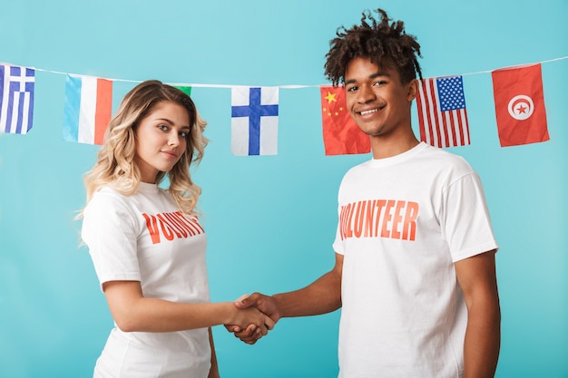 Heureux couple multiethnique confiant portant des volontaires t-shirt isolé sur mur bleu, se serrant la main