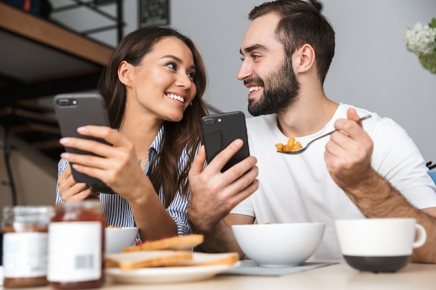Heureux couple multiethnique prenant son petit déjeuner dans la cuisine, à l'aide de téléphones mobiles