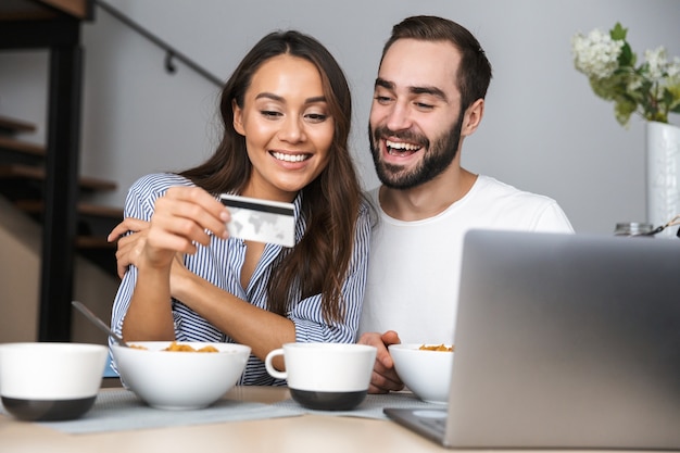 Heureux couple multiethnique prenant son petit déjeuner dans la cuisine, regardant un ordinateur portable, montrant la carte de crédit
