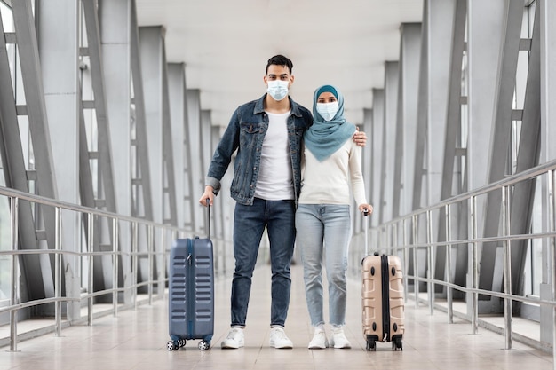 Heureux couple musulman dans des masques médicaux de protection posant avec des valises à l'aéroport