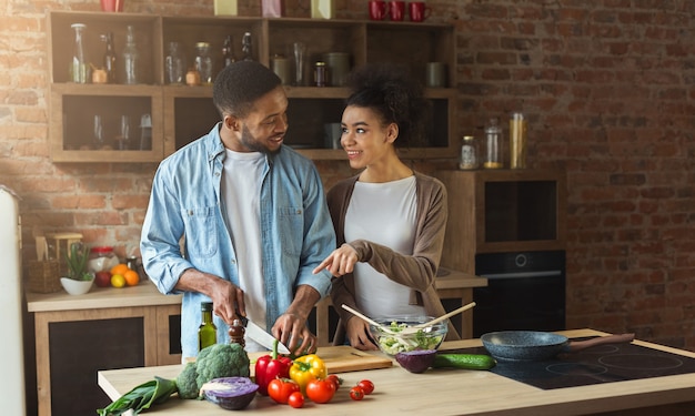 Heureux couple noir préparant une salade de légumes ensemble dans la cuisine loft. Dîner de cuisine de jeune famille