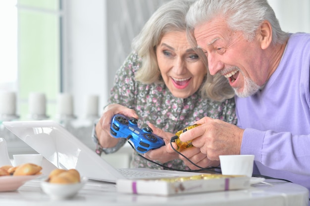 Heureux couple de personnes âgées jouant à un jeu d'ordinateur avec un ordinateur portable tout en buvant du thé dans la cuisine