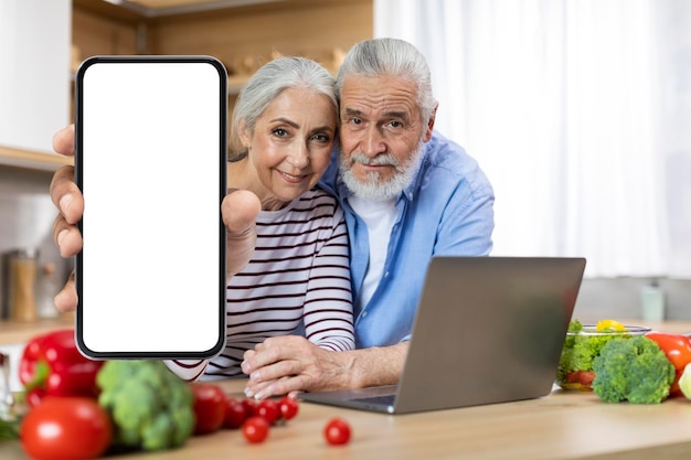 Heureux couple de personnes âgées montrant un grand téléphone portable vierge tout en posant dans la cuisine