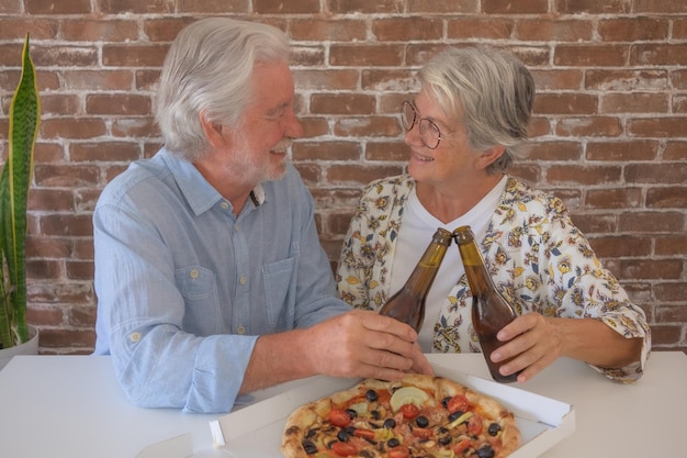 Heureux couple de personnes âgées portant un toast avec une bière se regardant Assis à la table de la maison prêt à manger une pizza Célébrer les vacances en compagnie