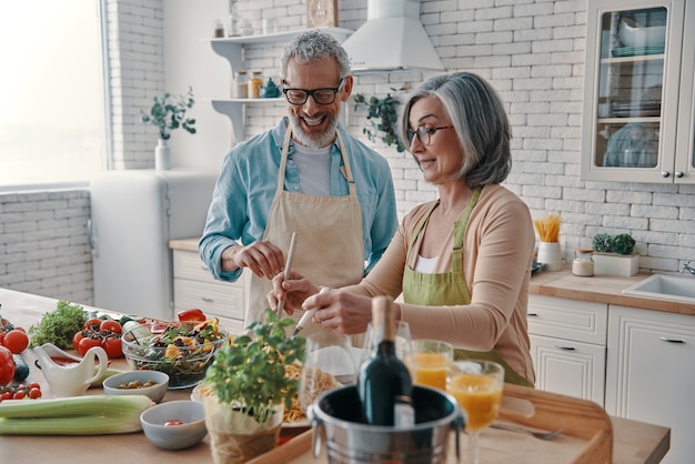 Heureux couple de personnes âgées en tabliers préparant un dîner sain et souriant tout en passant du temps à la maison