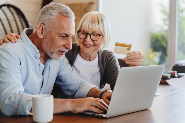 Heureux couple de personnes âgées utilisant un ordinateur portable et une carte de crédit pour faire des achats en ligne dans le salon à la maison