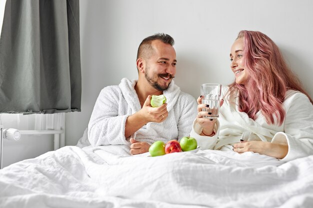 Heureux couple prendre le petit déjeuner sur le lit, ils rient en mangeant des pommes fraîches