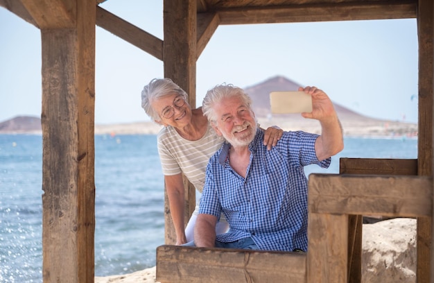 Heureux couple senior caucasien assis à l'ombre du belvédère devant la mer en utilisant le téléphone pour un selfie retraités âgés profitant de vacances à la mer par une journée venteuse et ensoleillée