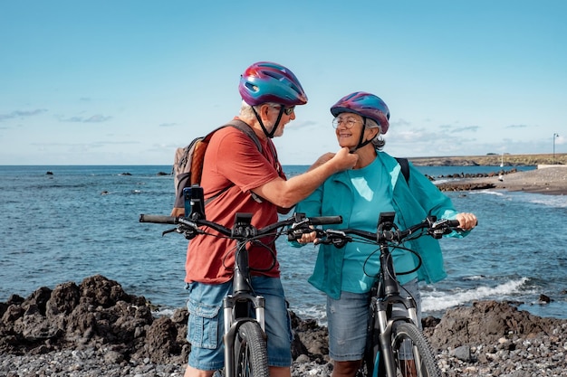 Heureux couple senior caucasien portant des casques à cheval sur la plage avec des vélos électriques Authentique concept de vie à la retraite pour personnes âgées Horizon sur la mer