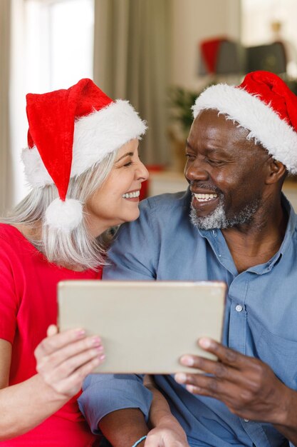 Photo heureux couple senior diversifié avec des chapeaux de père noël à l'aide d'une tablette. passer du temps de qualité à noël et au concept de retraite.