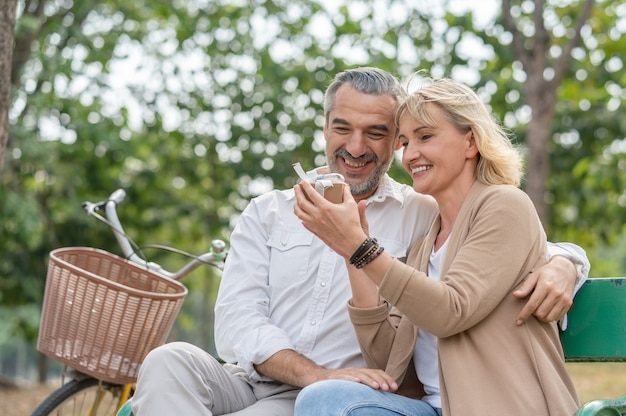 Heureux couple senior homme surprise donnant une boîte-cadeau à sa femme tout en vous relaxant et assis sur le banc dans le parc