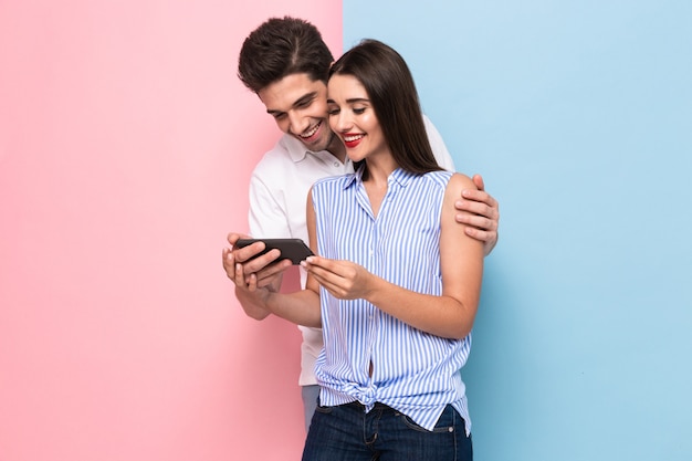 Heureux couple souriant et regardant le téléphone mobile, isolé sur un mur coloré