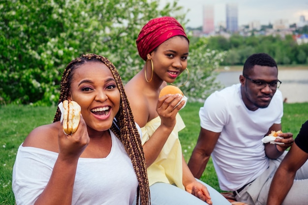 Heureux décontracté amérique africains s'amusant et mangeant des hamburgers à l'extérieur étudiants pour une pause soirée d'été temps nuageux dans le parc