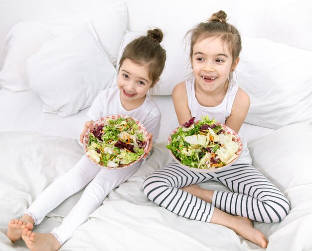 Heureux Deux Enfants Mignons Mangeant Une Salade De Légumes Dans La Chambre Sur Le Lit. Une Alimentation Saine Pour Les Enfants Et Les Adolescents.