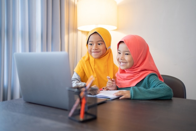 Heureux deux enfants musulmans portant un foulard souriant tout en utilisant un ordinateur portable à la maison
