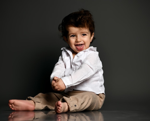 Heureux drôle flirter élégant bébé pieds nus portant un pantalon chemise montrant la langue Portrait isolé tourné sur fond gris studio Enfance heureuse et mode Croissance et développement des enfants