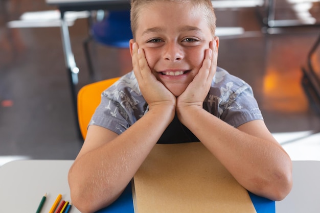 Heureux écolier caucasien assis au bureau dans la salle de classe s'appuyant sur des livres et souriant