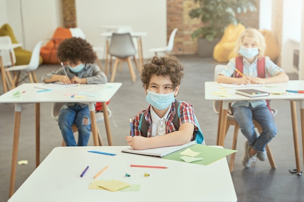 Heureux écolier portant un masque facial pendant l'épidémie de coronavirus souriant alors qu'il était assis au bureau à