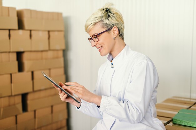 Heureux employé blond caucasien poursuivant tablette pour le travail tout en étant assis autour des boîtes dans l'usine alimentaire.