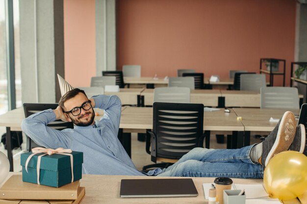 Photo heureux employé de bureau barbu positif en chapeau conique assis sur le lieu de travail avec ordinateur portable et cadeau avec ruban festif