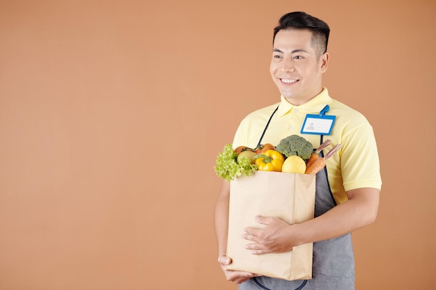 Heureux employé de magasin sombre asiatique portant un sac de produits frais collectés pour livraison au client