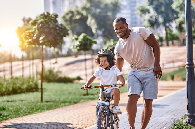 Heureux enfant afro-américain portant un casque tout en faisant du vélo et en faisant marcher son père à ses côtés