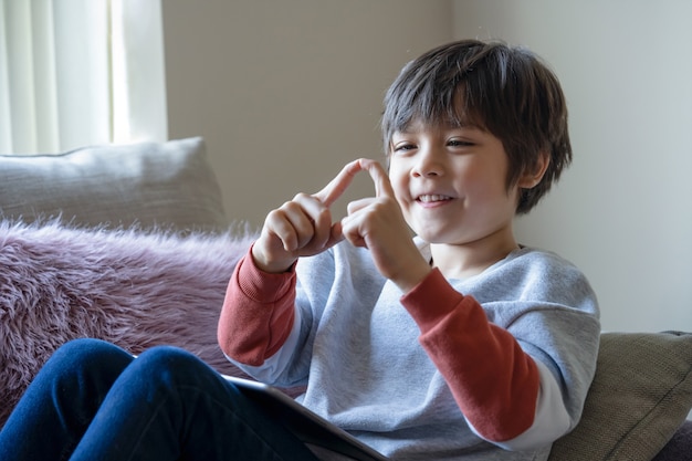 Heureux enfant assis sur un canapé en jouant avec ses doigts tout en regardant dessin animé sur tablette