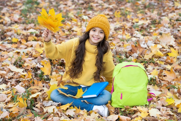 Heureux enfant au chapeau jaune tenir la feuille d'érable en plein air avec sac à dos scolaire se détendre dans le parc retour à l'école