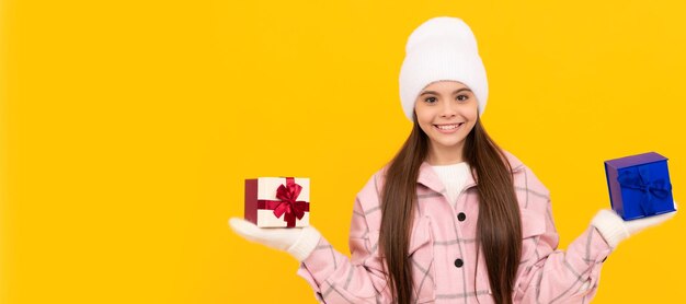 Heureux enfant en chapeau et mitaines choisir la boîte actuelle sur le choix de fond jaune Bannière de noël enfant fille studio enfant portrait d'hiver avec espace de copie