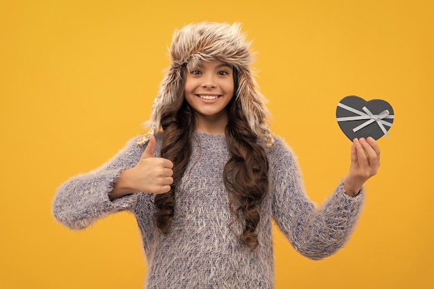 Heureux enfant cheveux longs en chapeau avec boîte-cadeau montrant le pouce vers le haut sur fond jaune saint valentin