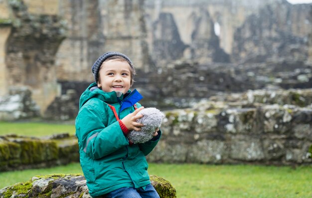 Heureux enfant garçon portant des chiffons chauds tenant sa peluche assis sur le vieux mur de briques avec des ruines floues de l'ancien fond de l'abbaye