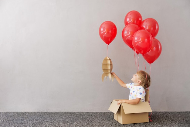 Heureux enfant jouant avec une fusée jouet à la maison. L'enfant fait semblant d'être astronaute. Garçon assis dans une boîte en carton avec des ballons rouges. Concept de rêve d'imagination et d'enfants