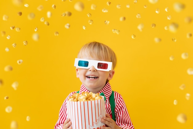 Heureux enfant portant des lunettes 3d et tenant des pop-corn contre le mur jaune.