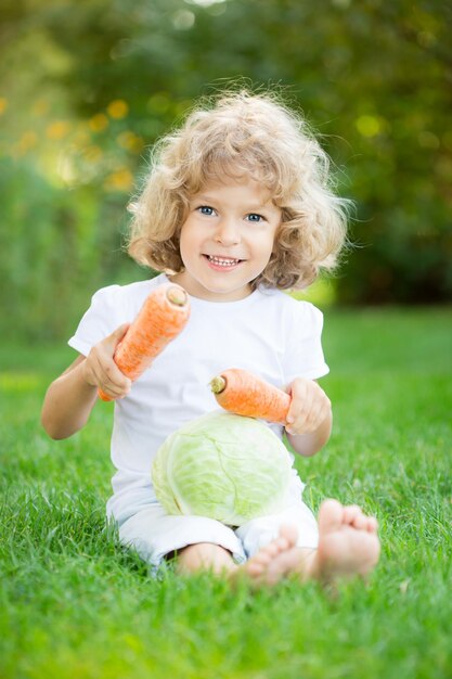 Heureux enfant souriant avec des légumes assis sur l'herbe verte dans le parc du printemps Concept de modes de vie sains